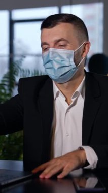 Takım elbiseli ve maskeli bir erkek, dizüstü bilgisayarın önünde oturuyor. Ofiste çalışırken salgın sınırlamalarını takip eden bir adam. Arka plan bulanık. Dikey video