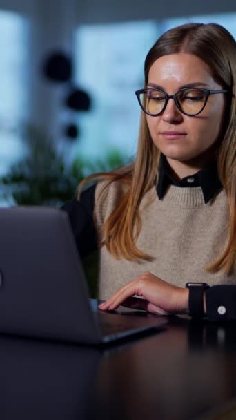 戴眼镜的金发女人打开她面前桌上的笔记本电脑 在办公室里从事计算机工作的年轻漂亮的女人 模糊的背景 垂直录像 — 图库视频影像