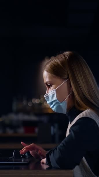 Stock video Young female employee using laptop. Woman wearing mask while working in the office. Side view. Blurred background. Vertical video