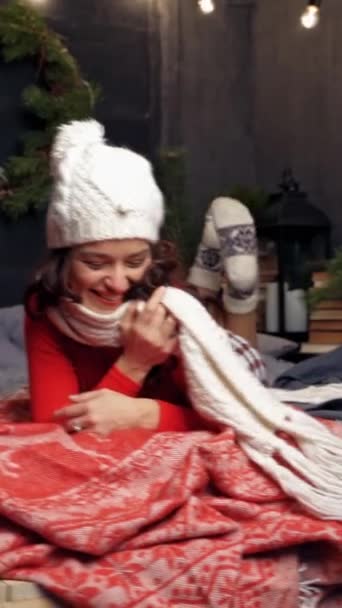クリスマスに感情的な女の子 編まれた帽子とスカーフで美しい若い女性は クリスマスのプレゼントやブランチの背景にベッドに横たわります バーティカルビデオ — ストック動画