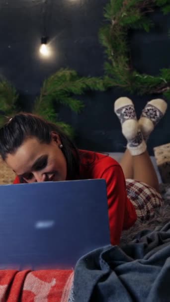 ベッドの上にラップトップを持っている魅力的な少女 美しい若い女性は 新しい年の木の近くでプレゼントとベッドに横たわっている間 ノートパソコンを見ています バーティカルビデオ — ストック動画