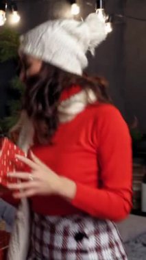 Yatakta hediye kutusunu fırlatan mutlu kız. Örgü şapkalı, elinde Noel hediyeleri olan ve evde eğlenen güzel genç bir kadın. Yeni yıl zamanı. Dikey video