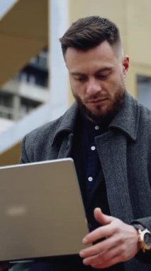 Laptoplu yakışıklı bir adamın portresi. Palto giyen genç sakallı iş adamı şehir arka planında kablosuz bir cihaz üzerinde çalışıyor. Dikey video