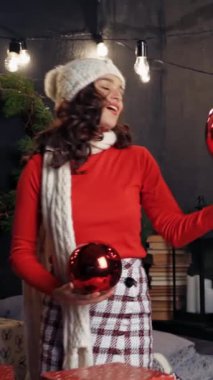Büyük kırmızı Noel topları olan güzel bir bayan. Beyaz örülü atkı ve şapkalı mutlu genç bir kadın yılbaşında güzel dekore edilmiş bir odada Noel mücevherleri bulundurur. Dikey video
