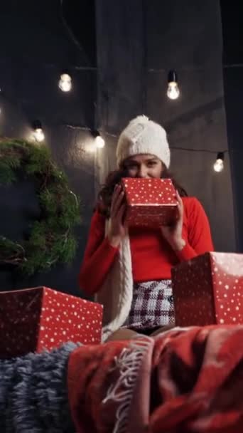 屋内でクリスマスプレゼントする美しい女の子 赤いセーターを着て幸せな若い女性の肖像画 ニットキャップとスカーフは自宅でクリスマスの背景に贈り物を投げます バーティカルビデオ — ストック動画