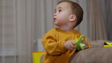 Sevimli beyaz çocuk kanepenin yanında elinde bir oyuncakla dikiliyor. Çocuk dikkatle yukarı bakar ve sonra kameraya döner..