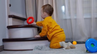 Turuncu spor takım elbiseli sevimli çocuk merdivenlerin yanında yerde oturuyor. Çocuk oyuncağı atar ve yuvarlanmasını izler..