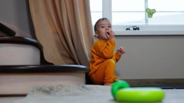 Pencerenin yanında oturan küçük bir çocuk ve içerideki merdivenler gözlerini ovuşturuyor. Bebek parmağını ağzına çeker ve odaya bakar..