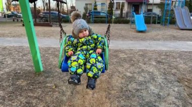 Sıcak giysiler ve şapkayla salıncakta oturan tatlı, ciddi bir erkek bebek. Çocuk dışarıda çocuk parkında vakit geçiriyor..
