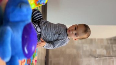 Dikey Ekran: Bir yaşındaki çocuk yerde oyuncaklarla oynuyor. Bebek çocuk oyuncaktan elini çırpar ve çevirir..