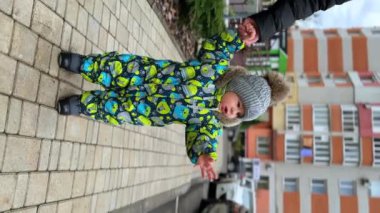 Dikey Ekran: Küçük bebek yürümeye başlıyor. Annenin eli oğlunu sıcacık elbiseler içinde dışarıda yürürken tutuyor..