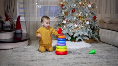 Turuncu spor takım elbiseli sevimli küçük çocuk, tatil için dekore edilmiş odada oyuncak piramit inşa ediyor. Arkaplanda Noel ağacı.