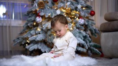 Beyaz elbiseli güzel bir çocuk Noel ağacının yanında pofuduk bir ekose üzerinde oturuyor. Çocuk bir oyuncak için heyecanlı. Kapat..