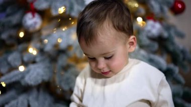 Güzel küçük çocuk Noel ağacı süslemesinin üzerinde oturuyor. Sevimli çocuk oyuncağın ve el sallamanın tadını çıkarıyor..