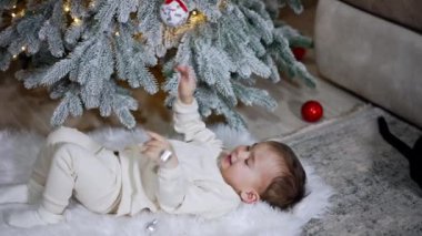 Beyaz elbiseli melek çocuk, Noel ağacının altında yumuşak bir ekose üzerinde yatıyor. Mutlu bebek bir oyuncakla oynuyor.