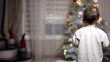 Odada yürüyen mutlu sevimli bebek. Çocuk kameraya tatlı tatlı gülümseyip el sallıyor. Arkaplanda Noel ağacı.