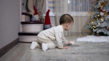 Beyaz elbiseli hoş beyaz çocuk Noel için dekore edilmiş odada oynuyor. Bebek kamerayı fark eder ve acele eder. Bulanık arkaplan.