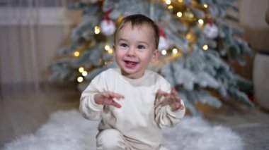 Gülümseyen mutlu beyaz çocuk heyecanla el sallıyor. Sevimli bebek Noel ağacının yanında yerde oturuyor..