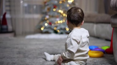 Sevimli bebek sırtüstü yere oturmuş Noel ağacına bakıyor. Çocuk hızla ağaca tırmanıyor..