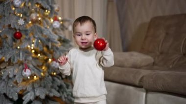 Sevimli çocuk Noel ağacından kırmızı oyuncağı aldığı için mutlu. Çocuk oyuncağı yere atar ve takip eder..