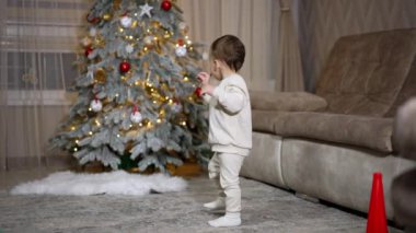 Beyaz elbiseli tatlı çocuk odada kameraya doğru duruyor. Bebek birkaç oyuncak almak için Noel ağacına yaklaşıyor..