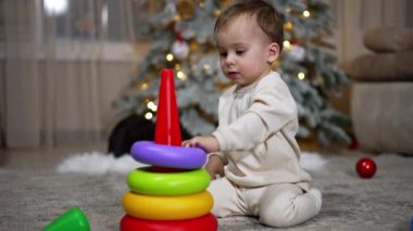 Beyaz elbiseli sevimli erkek bebek piramidi topluyor. Güzel bir Noel ağacı. Arkaplanda ışıklar var..