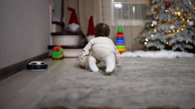 Beyaz elbiseli küçük çocuk odanın yanından geçip kamera ile takip ediyor. Bebek Noel ağacının yanındaki oyuncak piramidinin orada duruyor..