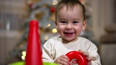 Küçük oğlan hapşırır ve mutlu bir şekilde güler. Çocuk ellerini sallayarak oyuncak piramitle oynuyor. Kapatın. Arkaplanda bulanık bir Noel ağacı.