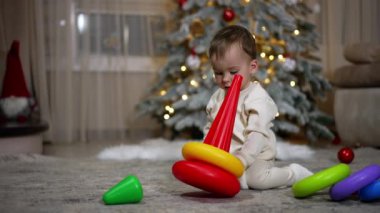 Kafkasyalı çocuk yerde oturup oyuncak piramitlerle oynuyordu. Dekorasyonlu Noel ağacı arka planda bulanık.