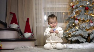 Tatlı beyaz çocuk, elinde emzik tutarak yerde oturuyor. Bebek kameraya doğru sürünüyor ve sonra Noel ağacına dönüyor..
