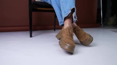 Büyük tabanlı kahverengi süet ayakkabılar ve aynı renk bağcıklar. Mavi kot giyen manken, kameranın önünde sandalyede oturup ayakkabılarını giyer. Kapat..