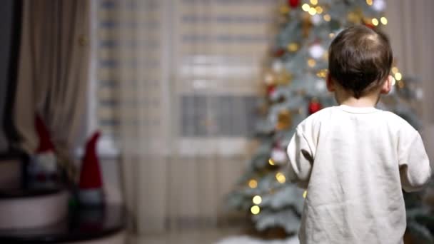 快乐可爱的婴儿在房间里走来走去 小孩对着镜头和挥手甜甜地笑着 背景下的圣诞树 — 图库视频影像