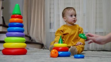 Turuncu takım elbiseli sevimli bebek oyuncaklarla çevrili oturuyor. Tatlı çocuk yana bakıyor ve çorabını ayağından çıkarıyor..