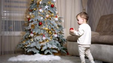 Sevimli bebeğin elinde Noel ağacından gelen kırmızı bir top var. Çocuk yaklaşan bir oyuncakla uzaklaşıyor..