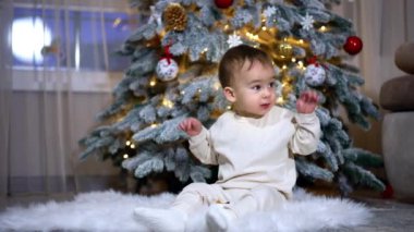 Beyaz elbiseli güzel melek gibi çocuk güzel dekore edilmiş Noel ağacının yanında oturuyor. Güzel çocuk bir oyuncakla oynar ve ellerini mutlu bir şekilde sallar..