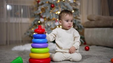 Oyuncak piramidin parçalarını toplayan mutlu çocuk. Bebek mutlu bir şekilde el sallıyor ve gülümsüyor. Kara kedi ön planda yürüyor. Arkaplanda Noel ağacı.