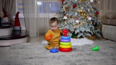 Sevimli bebek yüzükleri oyuncak piramidinden topluyor. Süslü Noel ağacıyla odada oynayan tatlı bir çocuk..