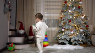 Bir yaşında bir çocuk elinde emzik tutarak odanın önünden geçiyor. Bebek güzel bir Noel ağacının yanından geçiyor pek aldırış etmiyor..