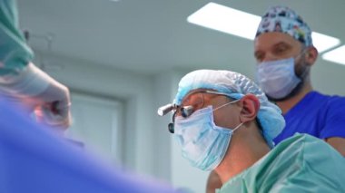 Maske, gözlük ve şapka takan cerrahın başı. Cerrah konuşurken doktor ameliyata odaklanıyor. Anestezi uzmanı. Düşük açı görünümü.