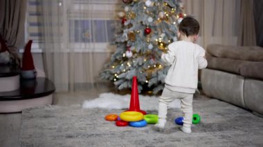 Beyaz elbiseli küçük bebek oyuncaklarına doğru yürüyor. Tatlı çocuk piramitlerle oynamak için yerde oturuyor. Arkaplanda Noel ağacı.