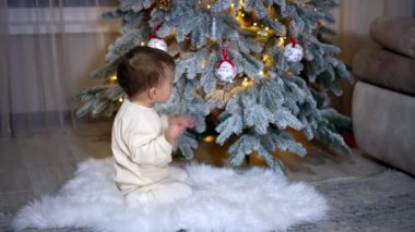 Süslü Noel ağacını kutlayan tatlı erkek bebek. Çocuk pofuduk beyaz ekose kumaşın üzerine oturur ve heyecanla el sallar..