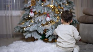 Tatlı bebek Noel ağacından kırmızı topu alıyor. Küçük çocuk tatil süslemelerinin ve kutlamaların tadını çıkarıyor..