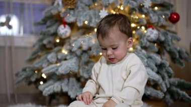 Güzel bebek halıya oturur ve oyuncağa odaklanır. Heyecanlı çocuk Noel ağacındaki oyuncaklarla oynuyor..