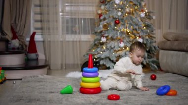 Noel ağacının yanında yerde oynayan ciddi, sakin bir bebek. Sevimli çocuk oyuncak piramidini topluyor..