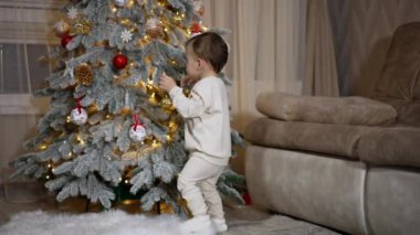 Beyaz elbiseli güzel çocuk Noel ağacından kırmızı top almak istiyor. Sevimli gülümseyen çocuk kameraya doğru döner..
