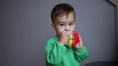 Koyu renk saçlı çocuk ağzına oyuncak dayıyor. Küçük bebek odada oyuncaklarla oynuyor..