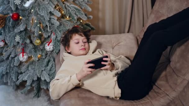 松懈的少年躺在沙发上 双脚向上 在电话上玩耍 孩子们的假日闲暇时间 背景下的圣诞树 — 图库视频影像
