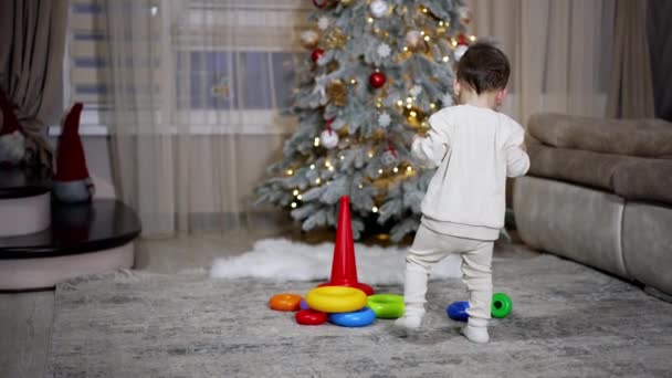 白い服を着た小さな赤ん坊は おもちゃまで歩いていく 美しい少年はピラミッドで遊ぶために床に座っています バックドロップのクリスマスツリー — ストック動画