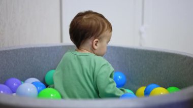 Beyaz tatlı çocuk kuru havuzun içinde oturuyor. Bebek bir top seçiyor, tutuyor ve kameraya bakıyor..