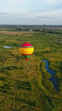 Renkli aerostat. Güzel sıcak hava balonu yeşil tarlaların arka planında ve kırsal alandaki küçük göllerde havada seyahat eder. Hava görüntüsü. Dikey video